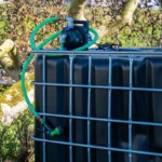 cuve-ibc-noir-avec-pompe-220V-1000L-renouvele-palette-acier-plastique-zijaanzicht-achteraanzicht-met-uitgaande-slang-in-tuinomgeving.webp