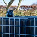 cuve-ibc-noir-avec-pompe-220V-1000L-renouvele-palette-acier-plastique-zijaanzicht-achteraanzicht-zonder-uitgaande-slang-in-tuinomgeving.webp