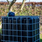 cuve-ibc-noir-avec-pompe-sur-batterie-1000L-renouvele-palette-acier-plastique-vue-de-cote-arrieere-sans-tuyau-sortant-dans-environnement-de-jardin.webp